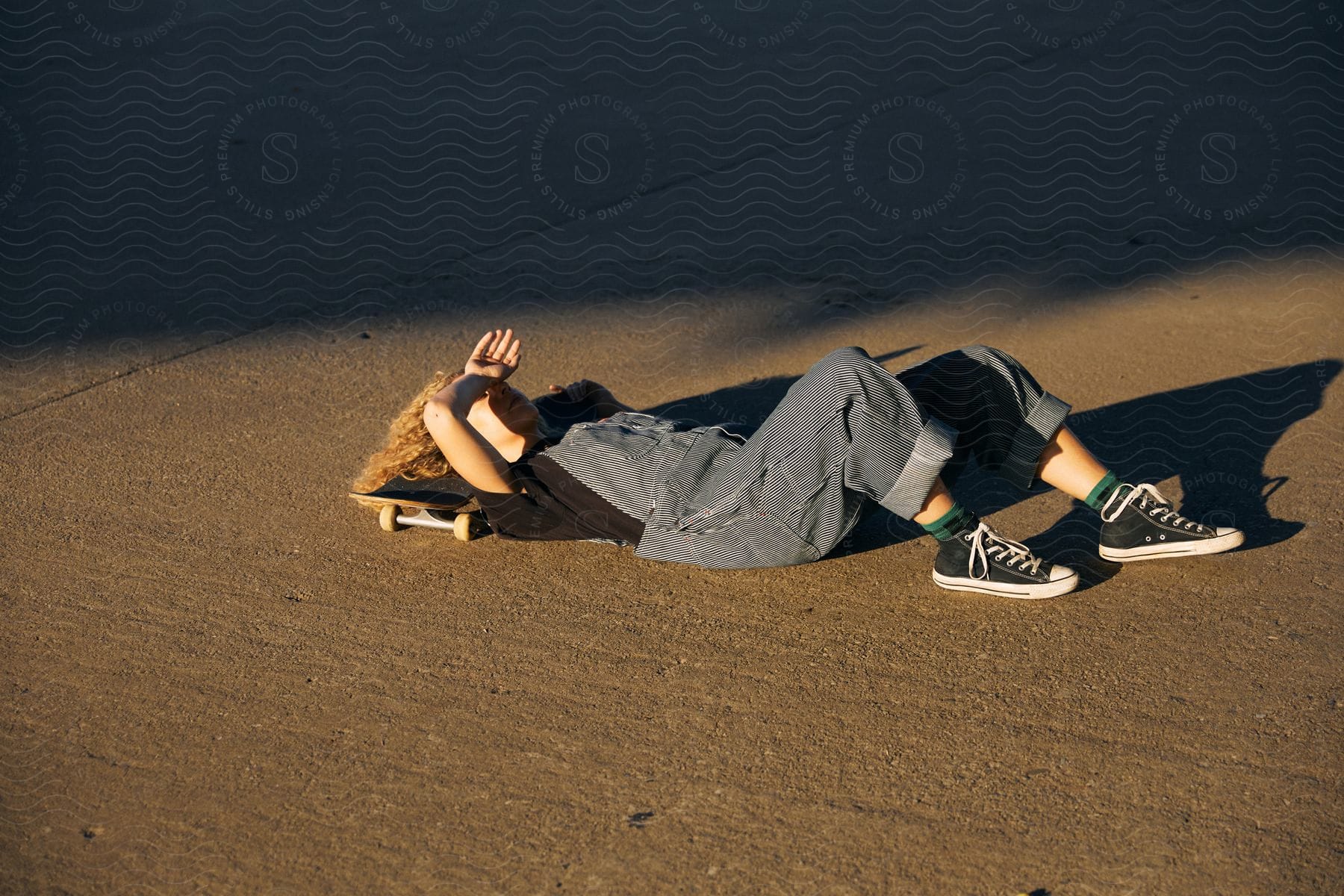 A skateboarder in overalls, green socks, and Converse shoes, with blond fizzly hair, rests on the asphalt with their head on their board, shielding their eyes from the sun.