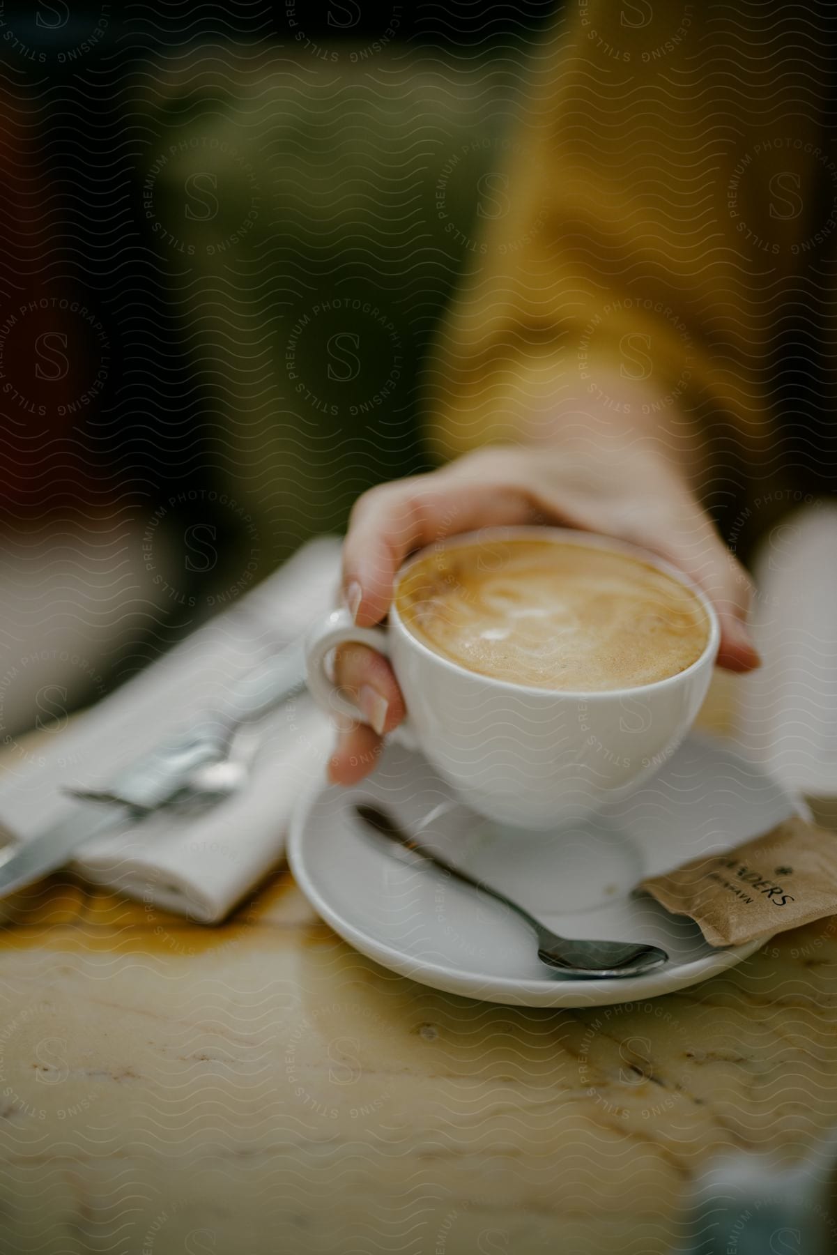 A person holds a cup of coffee in one hand, the saucer in the other, a spoon resting on it.