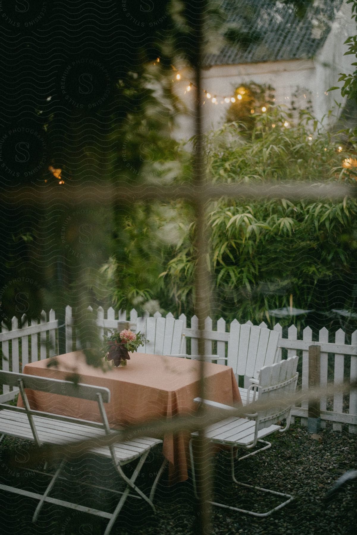 View out of a window of a bench and chair pulled up to a table with a table cloth and flowers in a vase inside a white picked fence in a neighborhood backyard