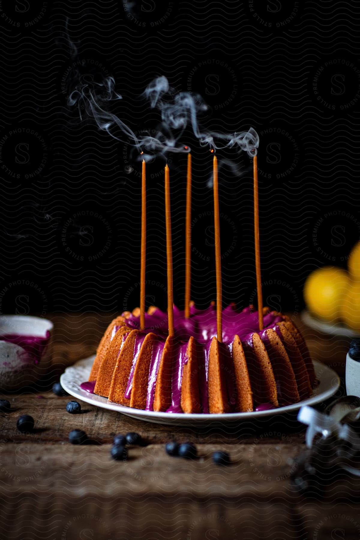 A blueberry glazed bunt cake with candles is on a plate with blueberries on a table with a cup and plate of oranges