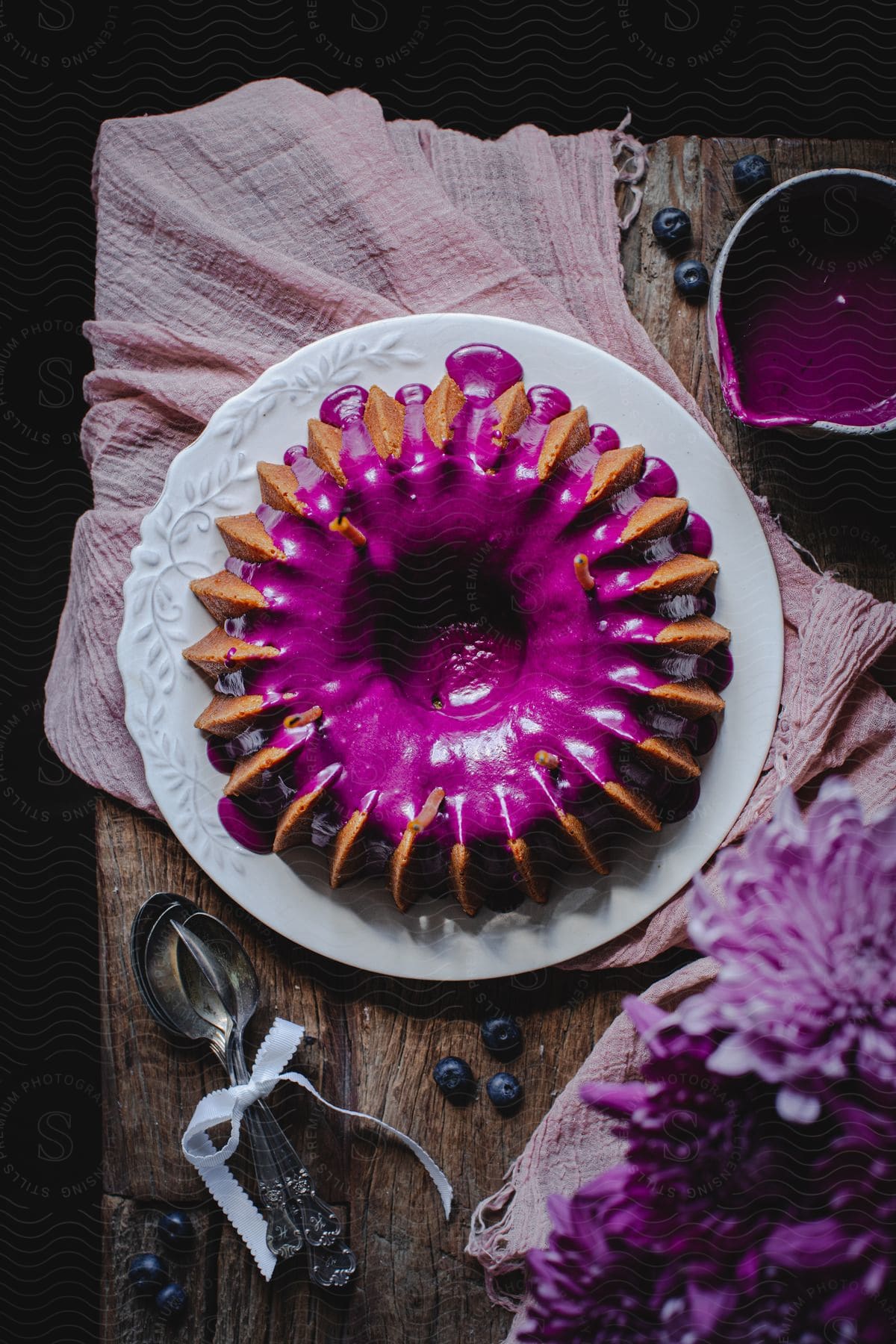 A purple-iced bundt cake on a white plate, surrounded by purple flowers, blueberries, and purple frosting, sits on a wooden tablecloth.