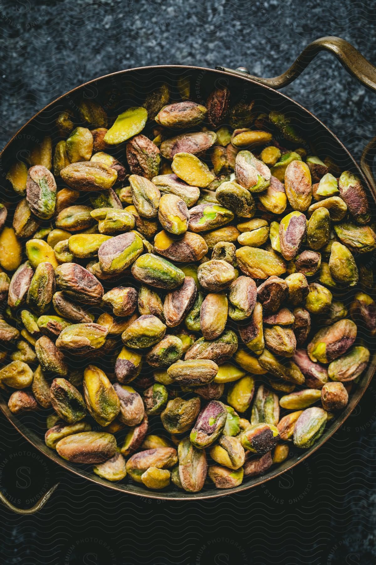 A pan of roasted pistachios, their shells discarded, rests on a cool granite countertop.