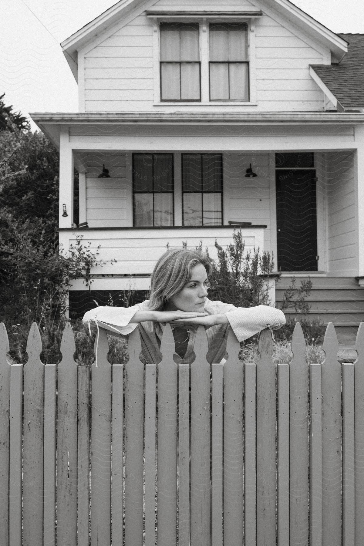 A woman standing in her front yard chin resting on her arms on top of a picket fence