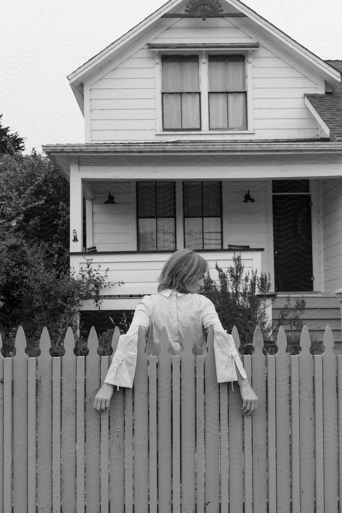 A woman is standing in front of her house with her back leaning against the fence and her arms over the fence