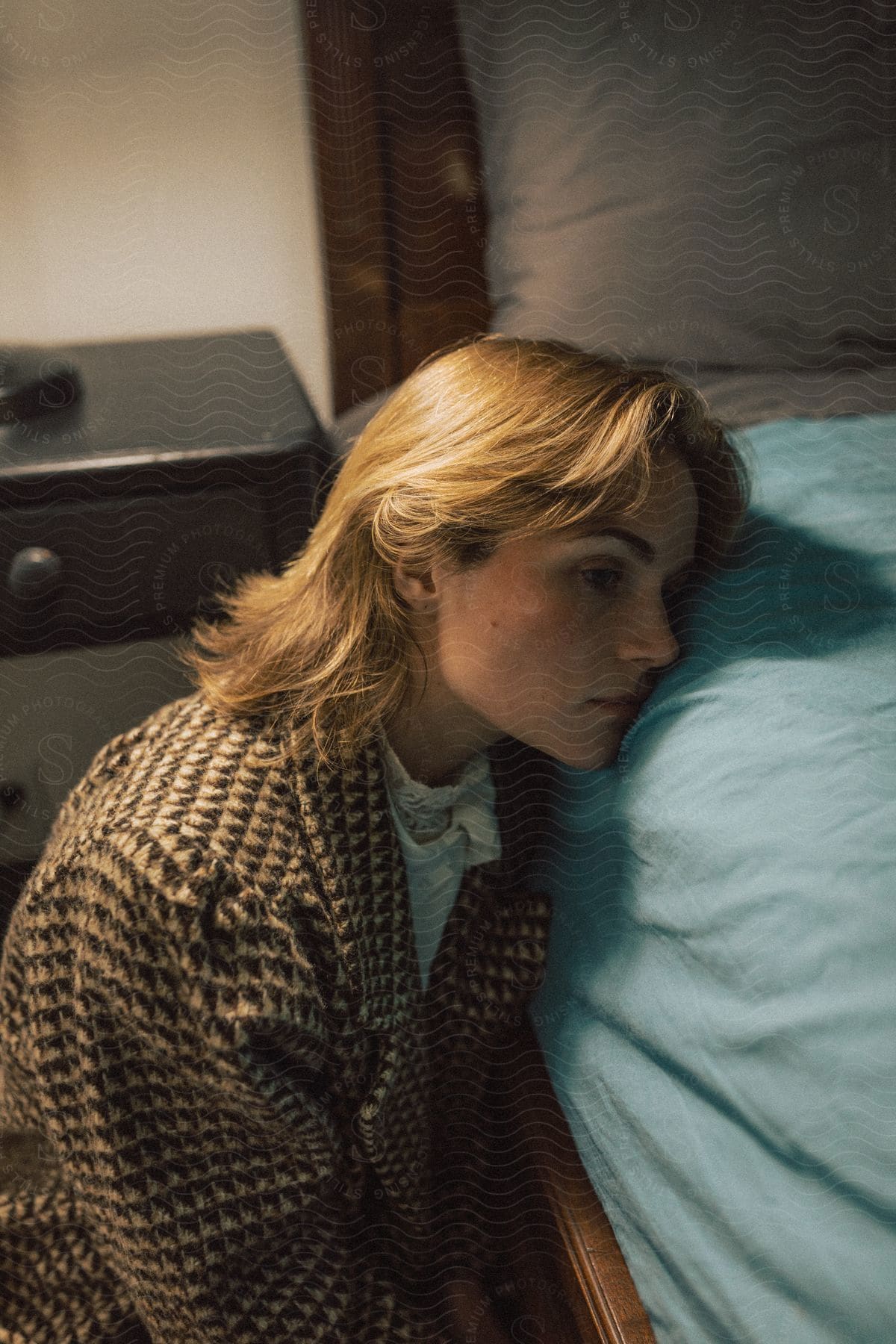 A blonde woman with sad facial expression sitting beside her bed.