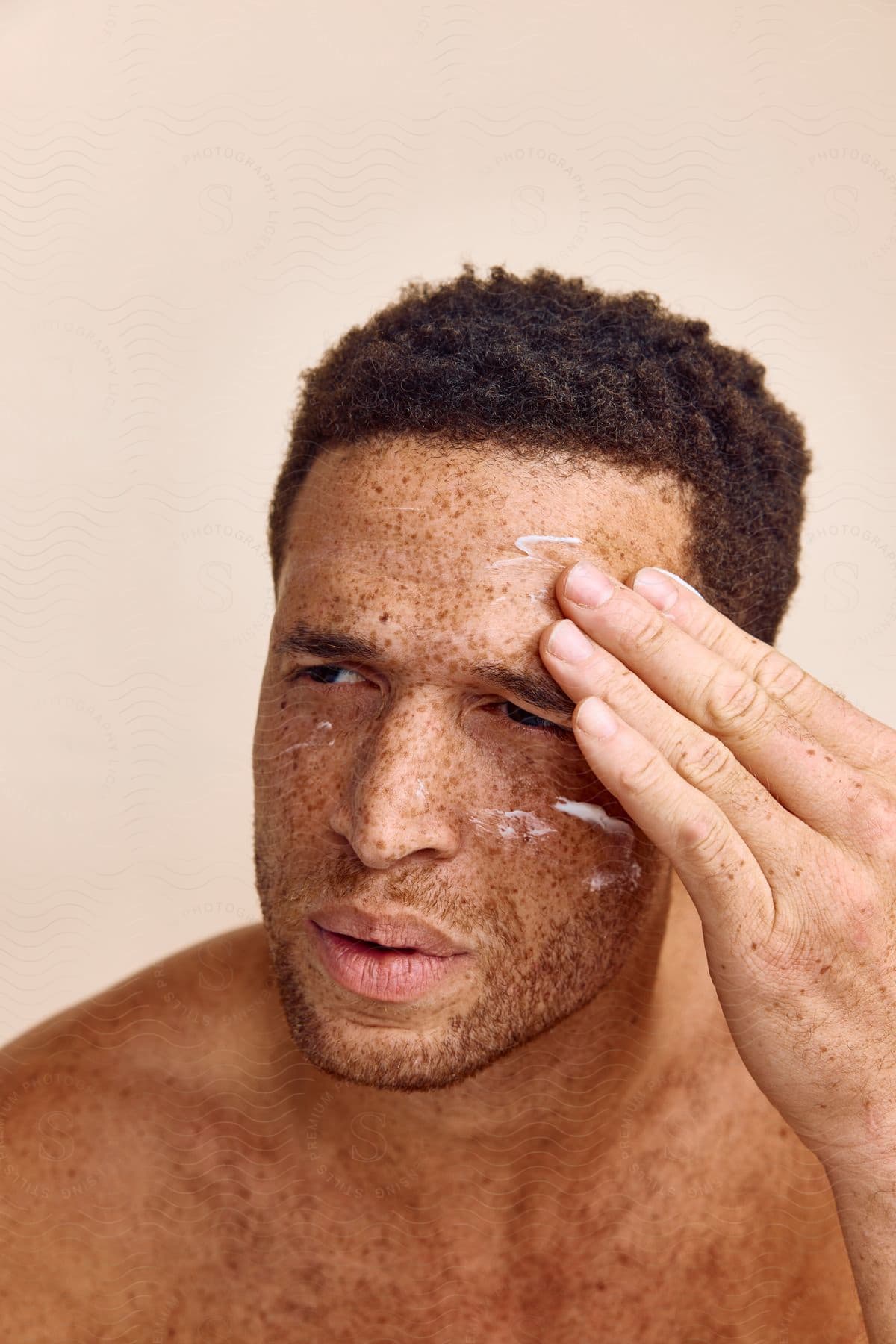 A shirtless freckled man applying face cream.
