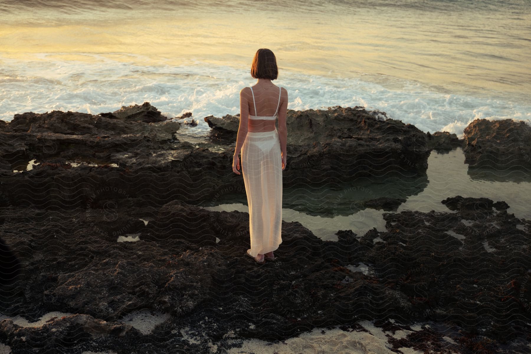 A woman wearing a see-through white dress is standing on a rocky beach looking at the ocean waves.