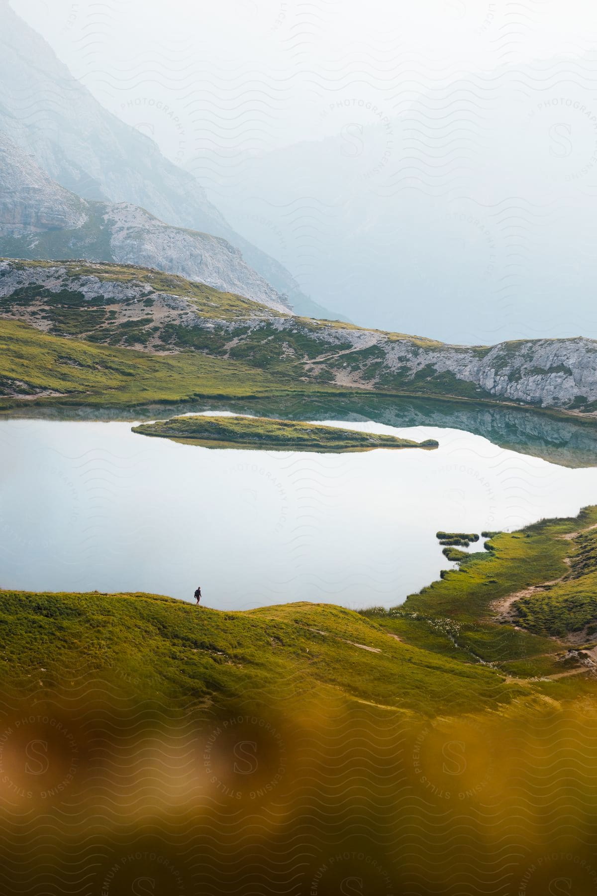 A person is walking on a path near the body of water that is below the mountain.