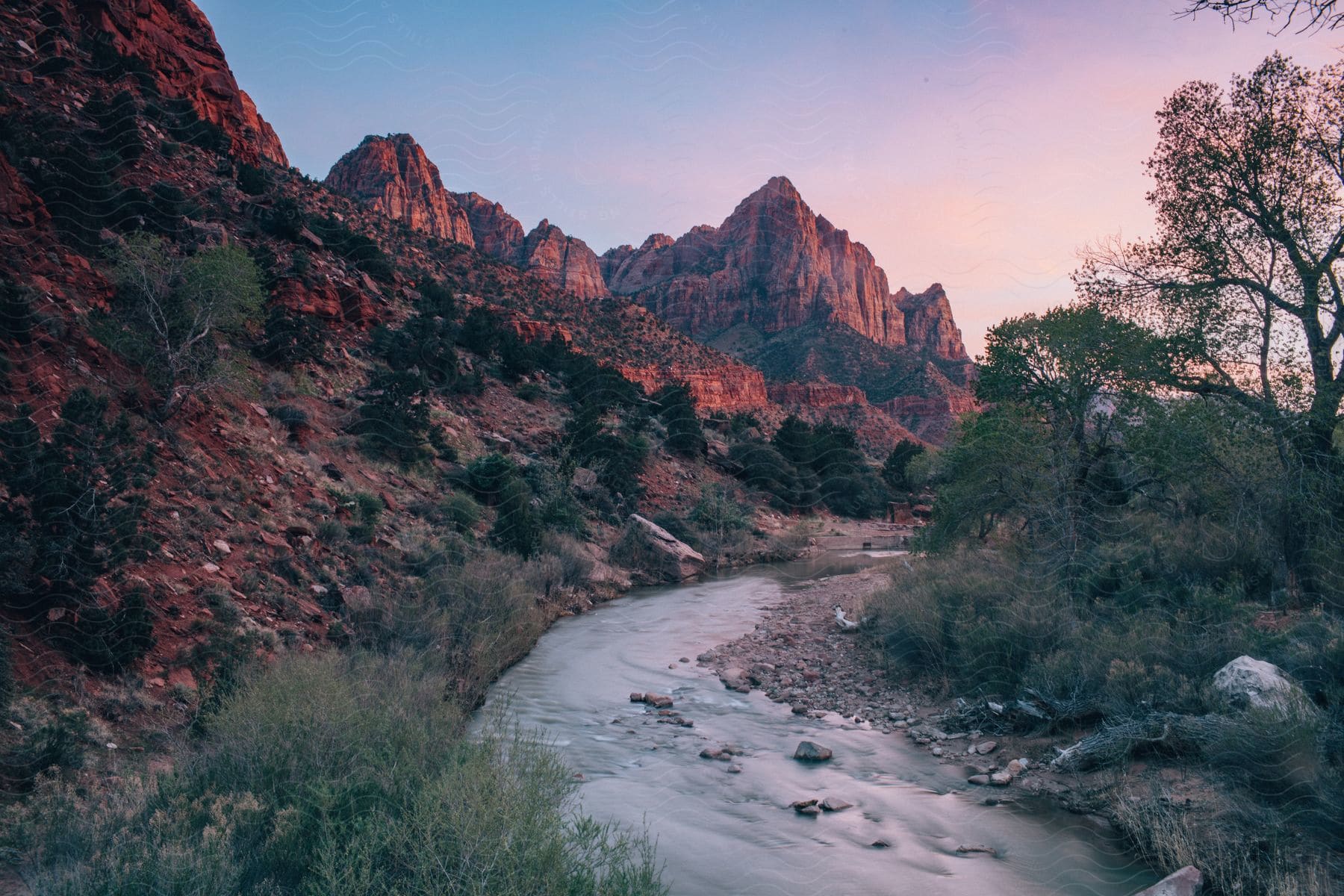 A river winds through a forest, reflecting the setting sun on the mountains to the left.