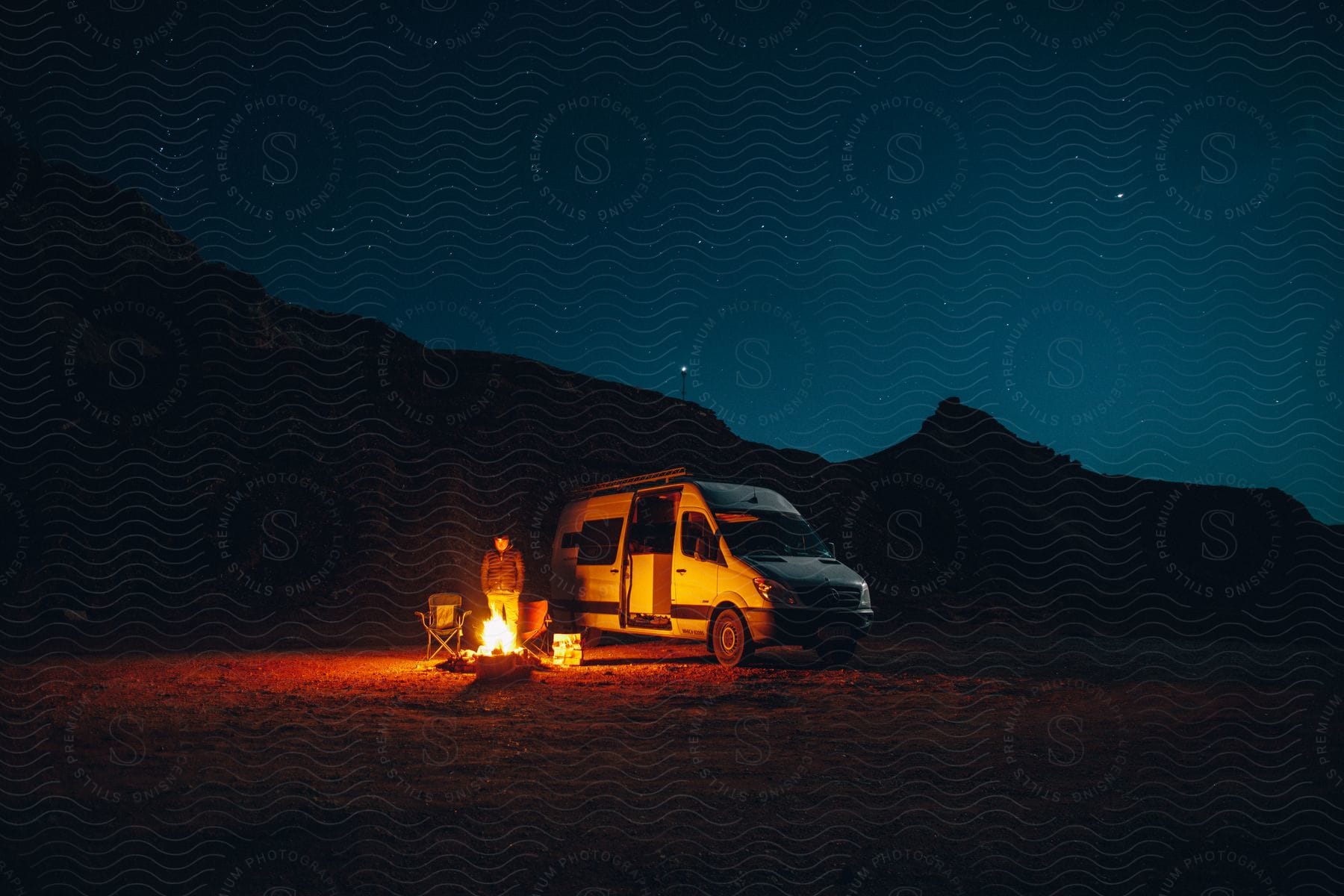 Man stands by campfire near van parked close to silhouetted mountains at night.