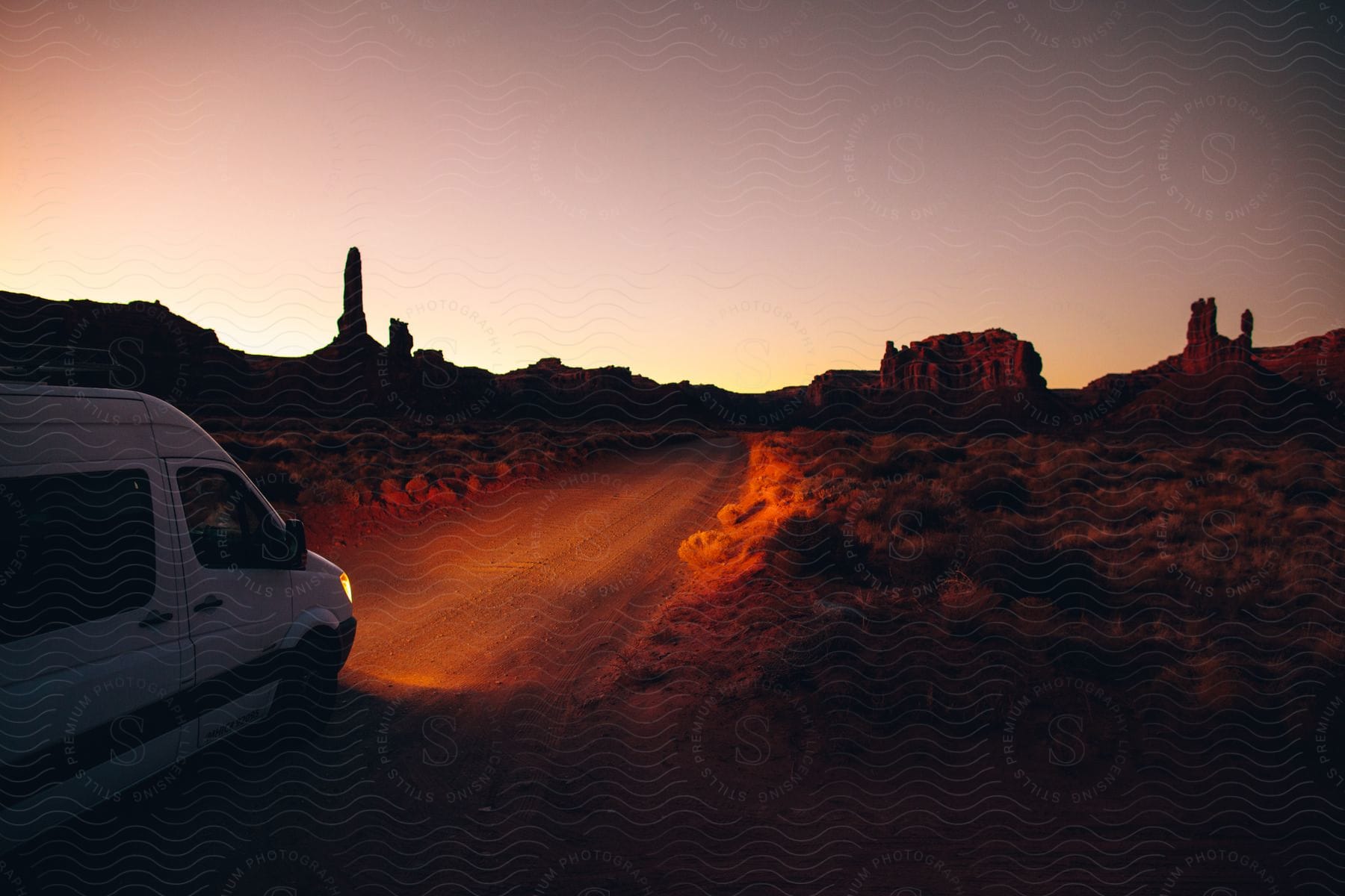 Van headlights illuminate a dirt road through a desert landscape, with a rock formation silhouetted at sunset.