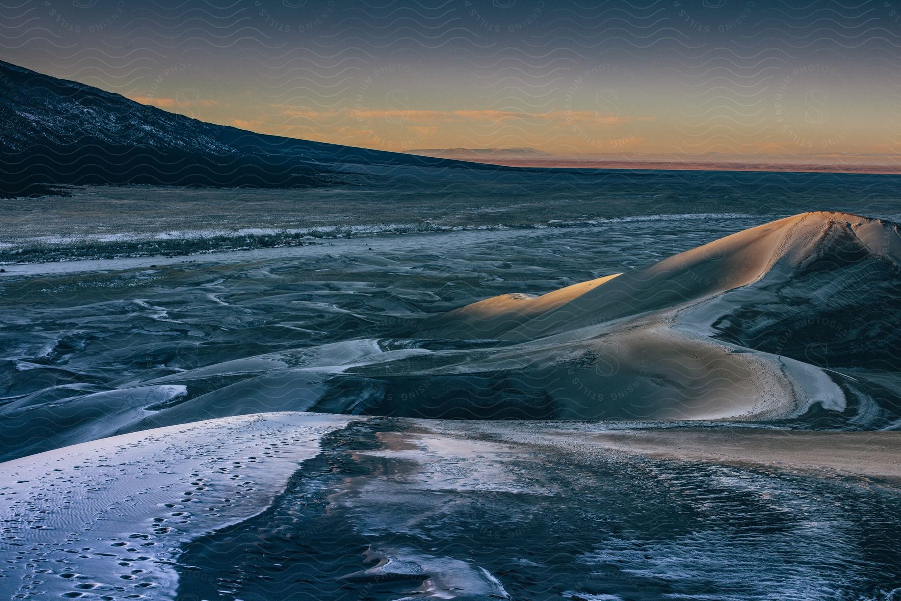 Ice and snow cover water and flatland near base of slope under a red tinted sky.