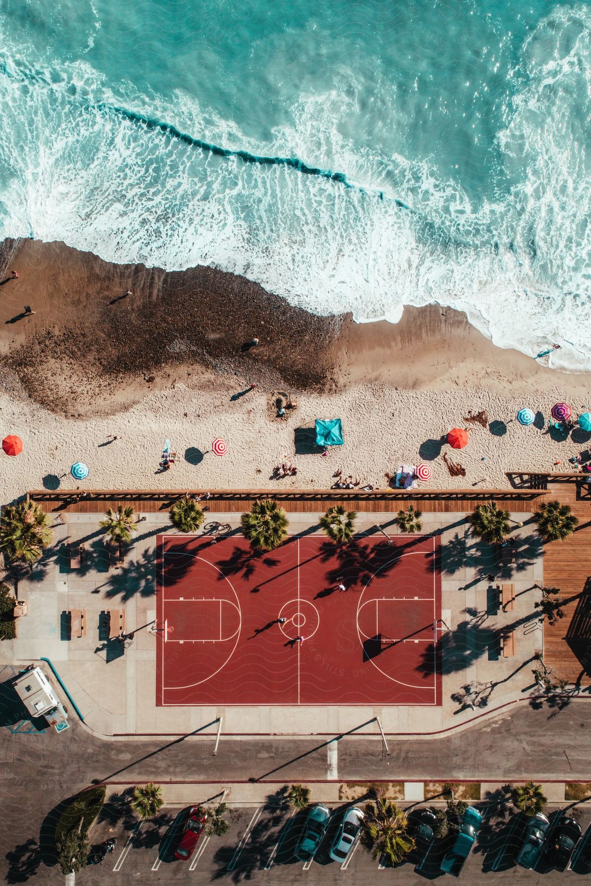 Aerial view of a beach basketball court with people playing, sunbathers under umbrellas, and waves crashing on the shore.