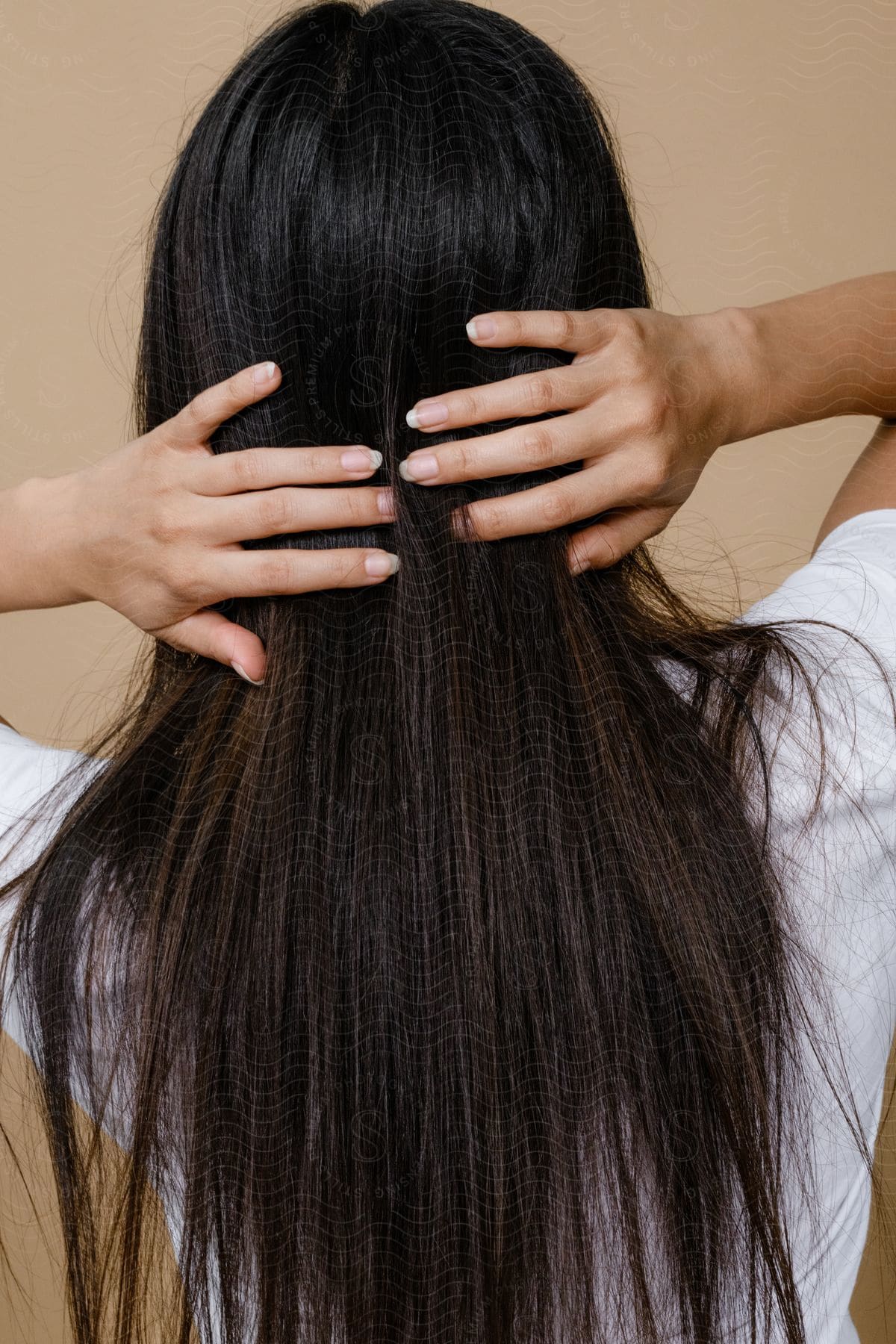 A woman with long dark brown hair is standing with her back turned and her hands on her hair on the back of her head