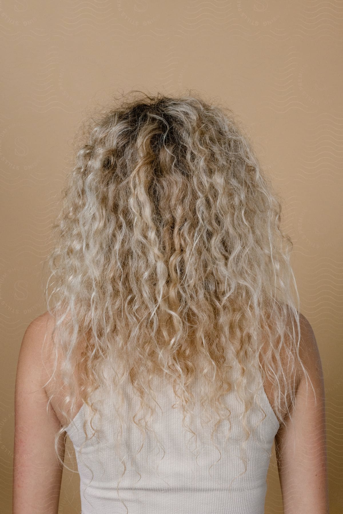 A blonde woman with curly hair in a white tank top stands before a wall.