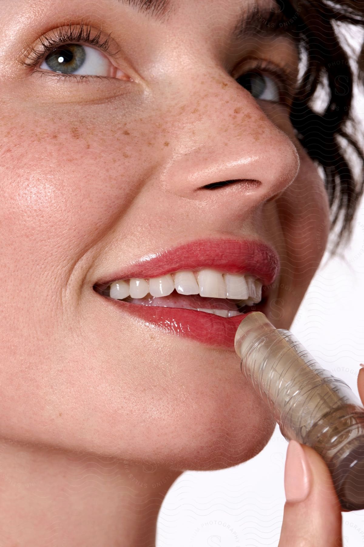 A freckled woman with red lips smiles while applying clear lip gloss.