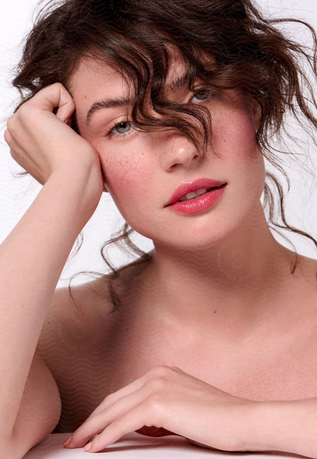 A woman with rose-hued cheeks and tousled hair strikes a pose with her hands on her head for a portrait.