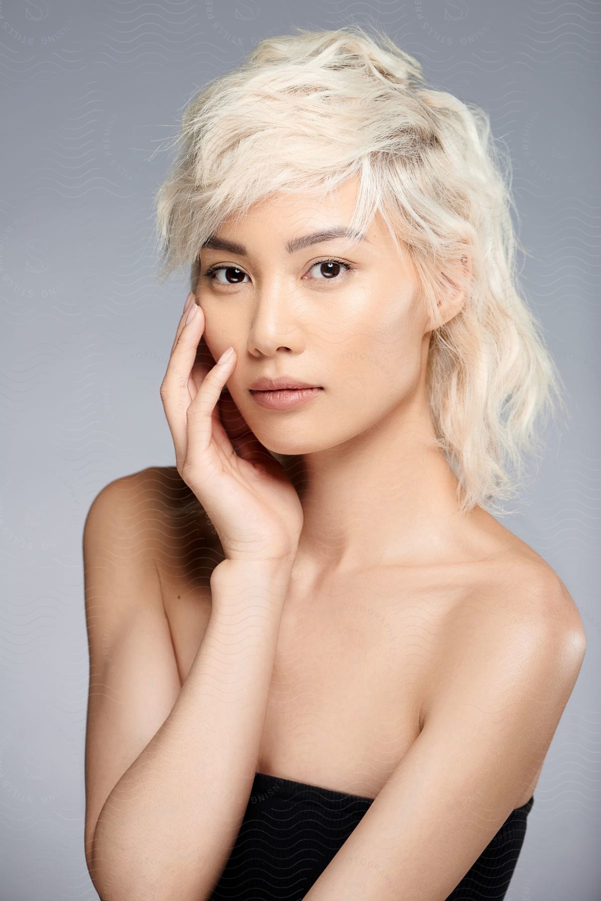 A bare shouldered blonde haired woman is looking ahead with her hand on her face