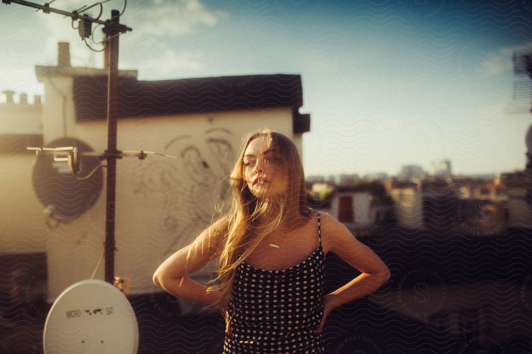 A woman in a polka dot dress, her hair blowing in the wind, stands on a rooftop in front of a satellite dish, overlooking the city.