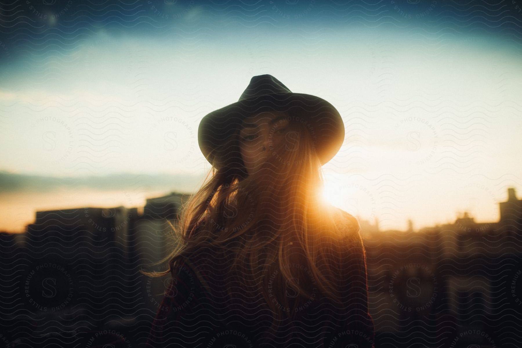 A portrait of a young woman wearing a hat with the sun setting on the horizon.