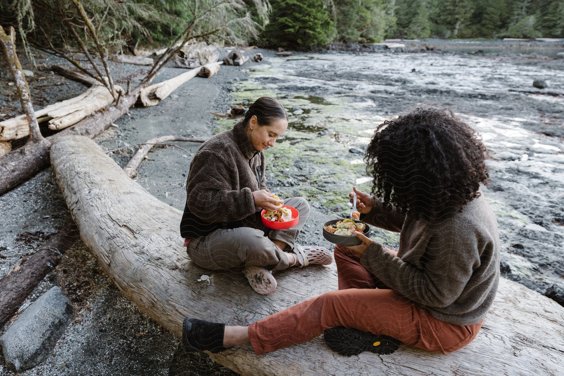 Two women sitting on a wooden log by the river, having a meal.