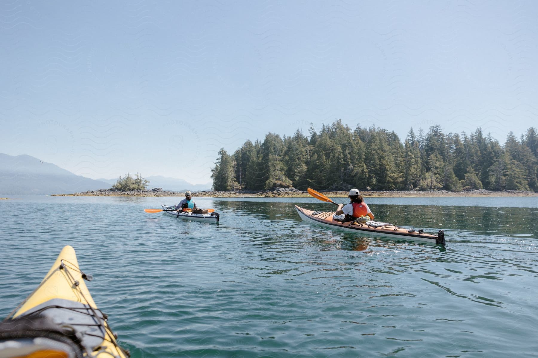 Two people canoeing on a tranquil lake, surrounded by trees and blue skies, with a glimpse of another canoe in the foreground.