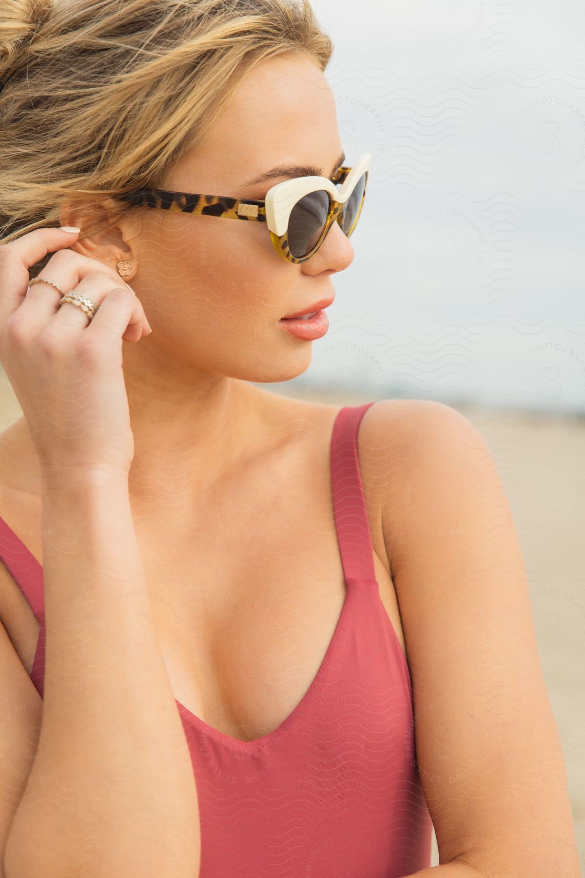 A woman wearing sunglasses and a swimsuit is on the beach with her head turned and her hand on her ear