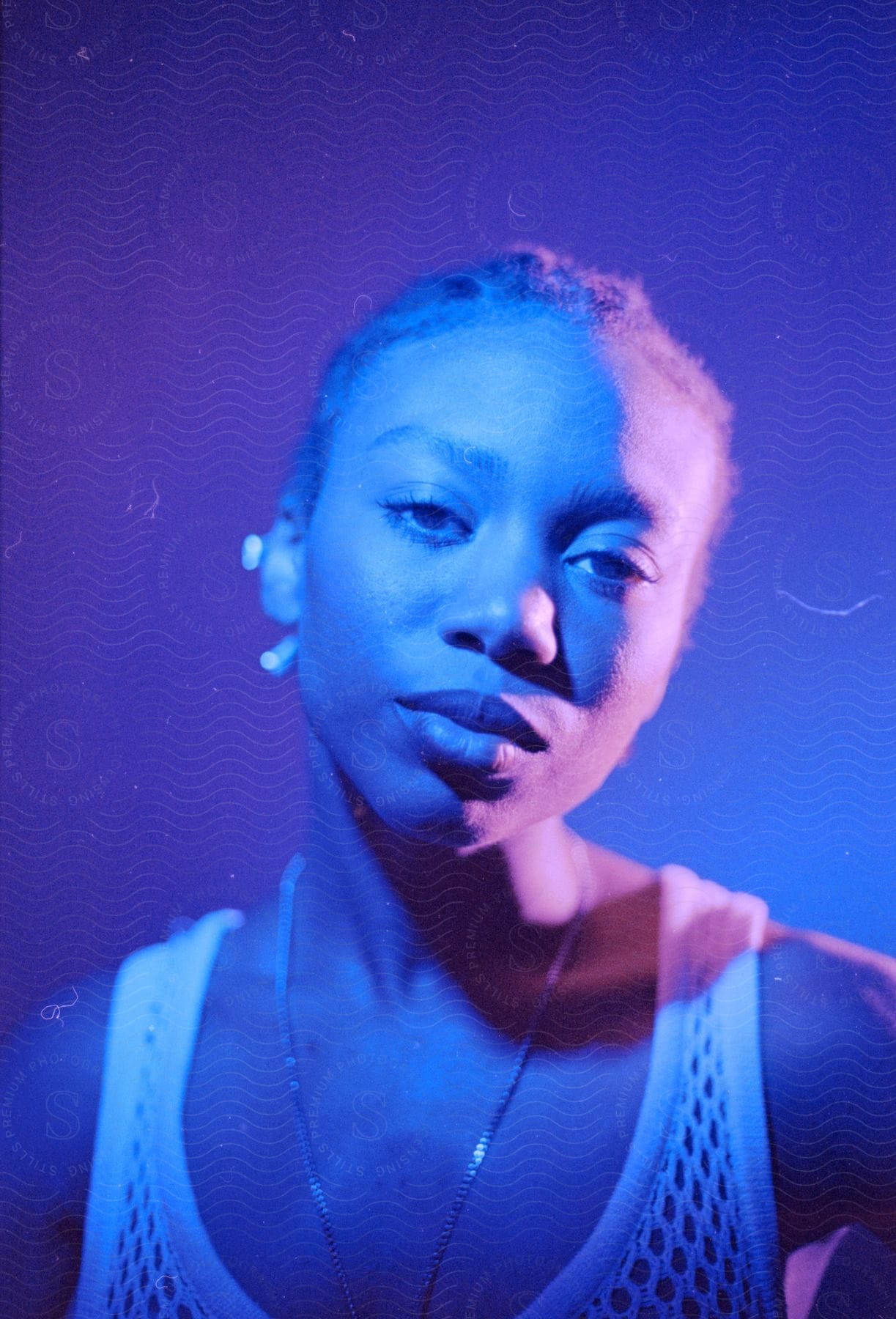 An African-American woman in a white tank top poses in blue light with a purple backdrop, a chain necklace adorning her neck.
