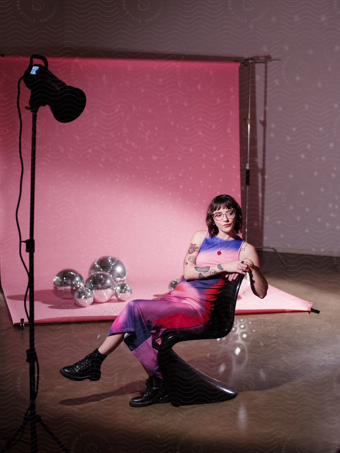 A woman in a full-body multicolored dress sits in a black chair in a studio with a pink backdrop and spotlight, silver balls of various sizes at the base of the backdrop.