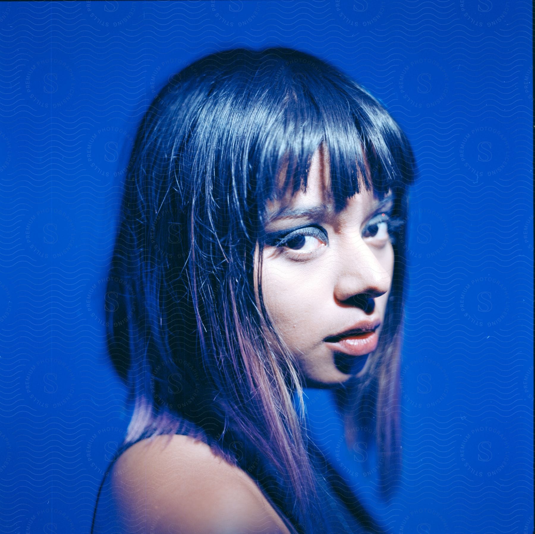 A woman with shoulder-length black hair and blue eyeshadow poses for a picture against a blue background with white light illuminating her face.