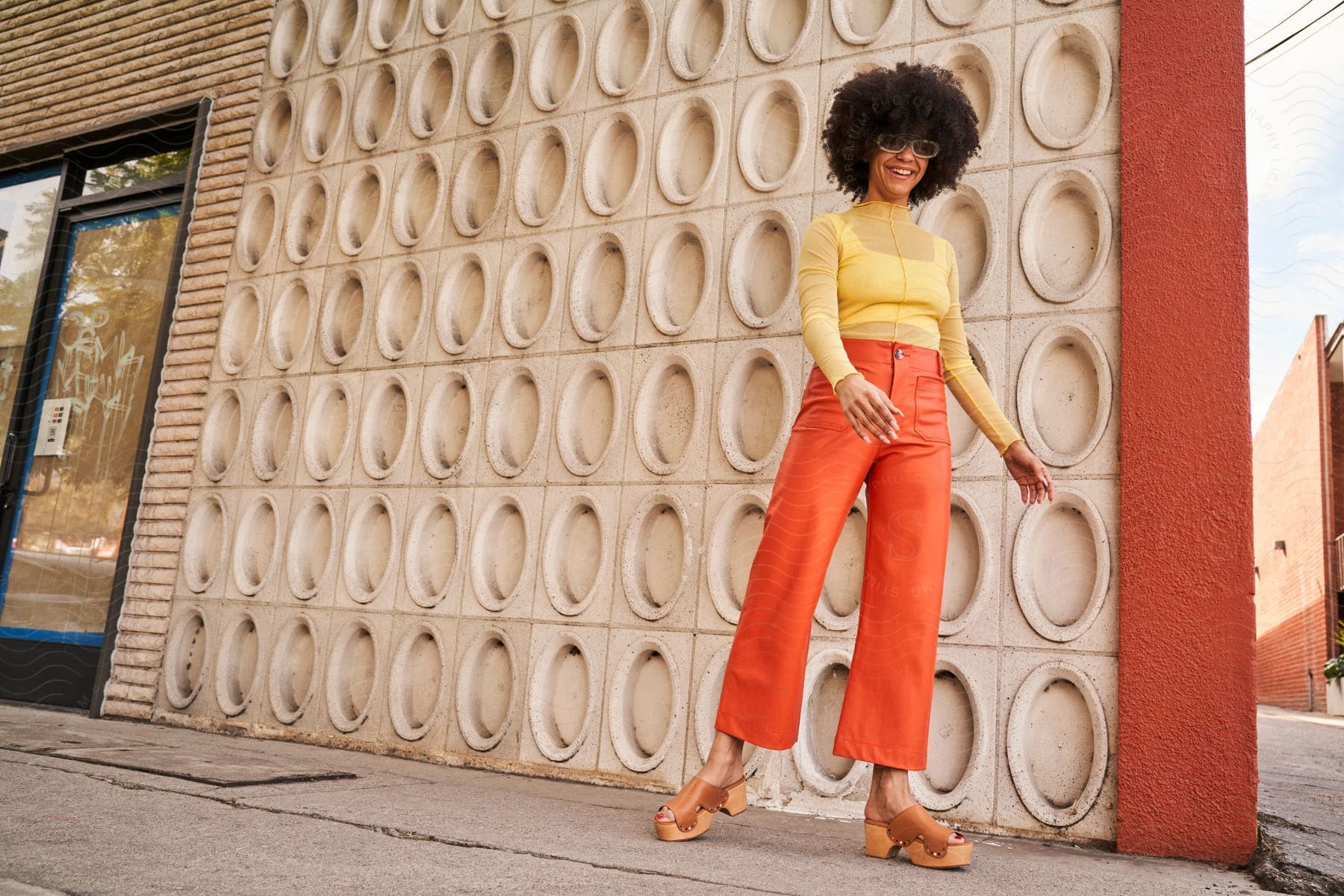 Woman with frizzy hair sunglasses yellow shirt and orange pants laughs in front of building’s wall