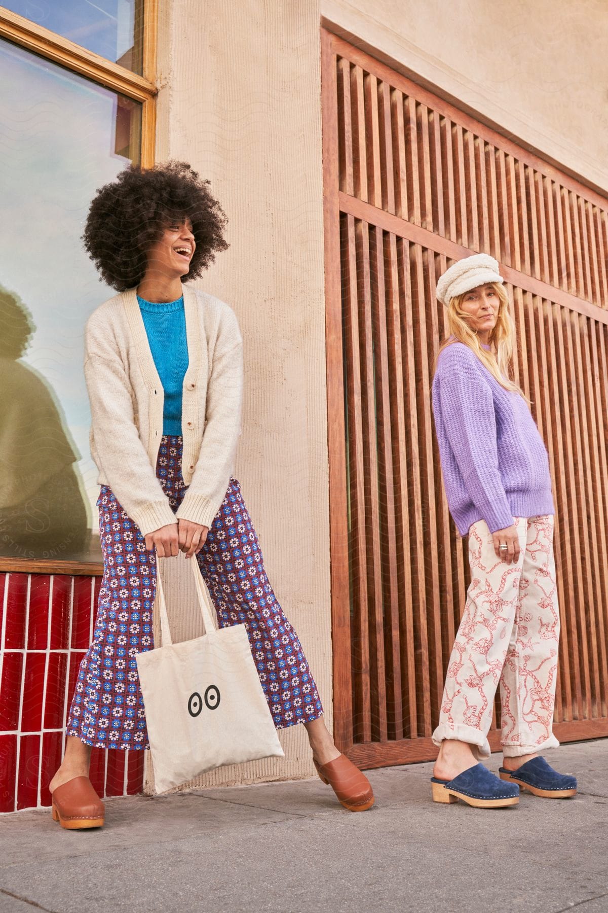 Two woman are modeling early spring fashion while outside next to a building in the daytime.