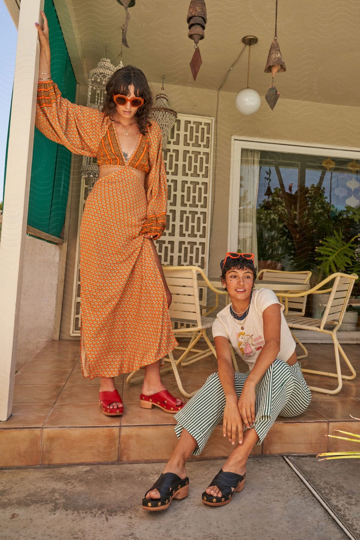 Two women on a patio, one standing on the patio in an orange patterned dress and red shoes, the other sitting on the patio step in a white shirt with a cupid character, blue striped pants, black shoes, and orange sunglasses.