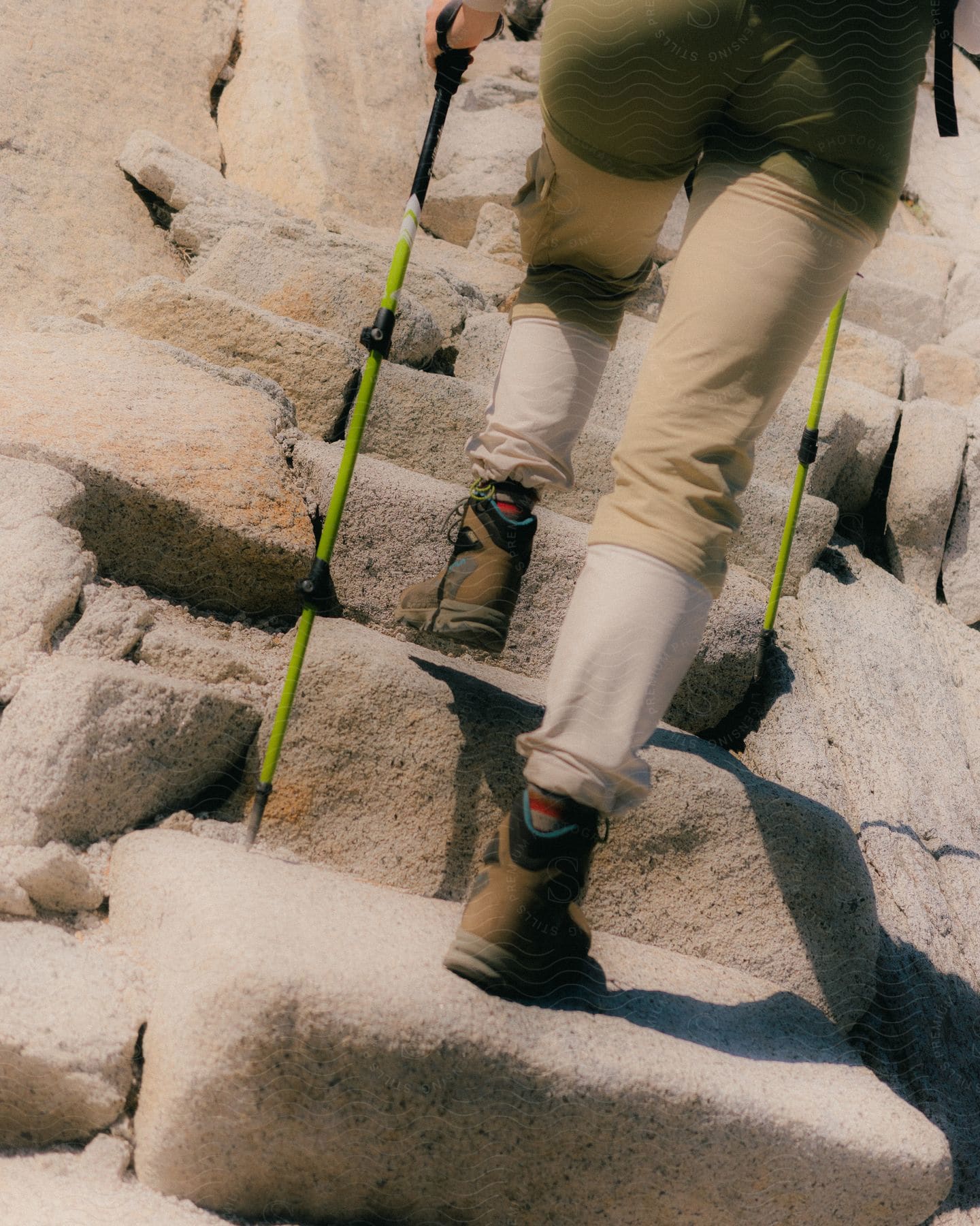 A hiker in hiking boots and cargo pants ascends rocky terrain with green hiking poles.