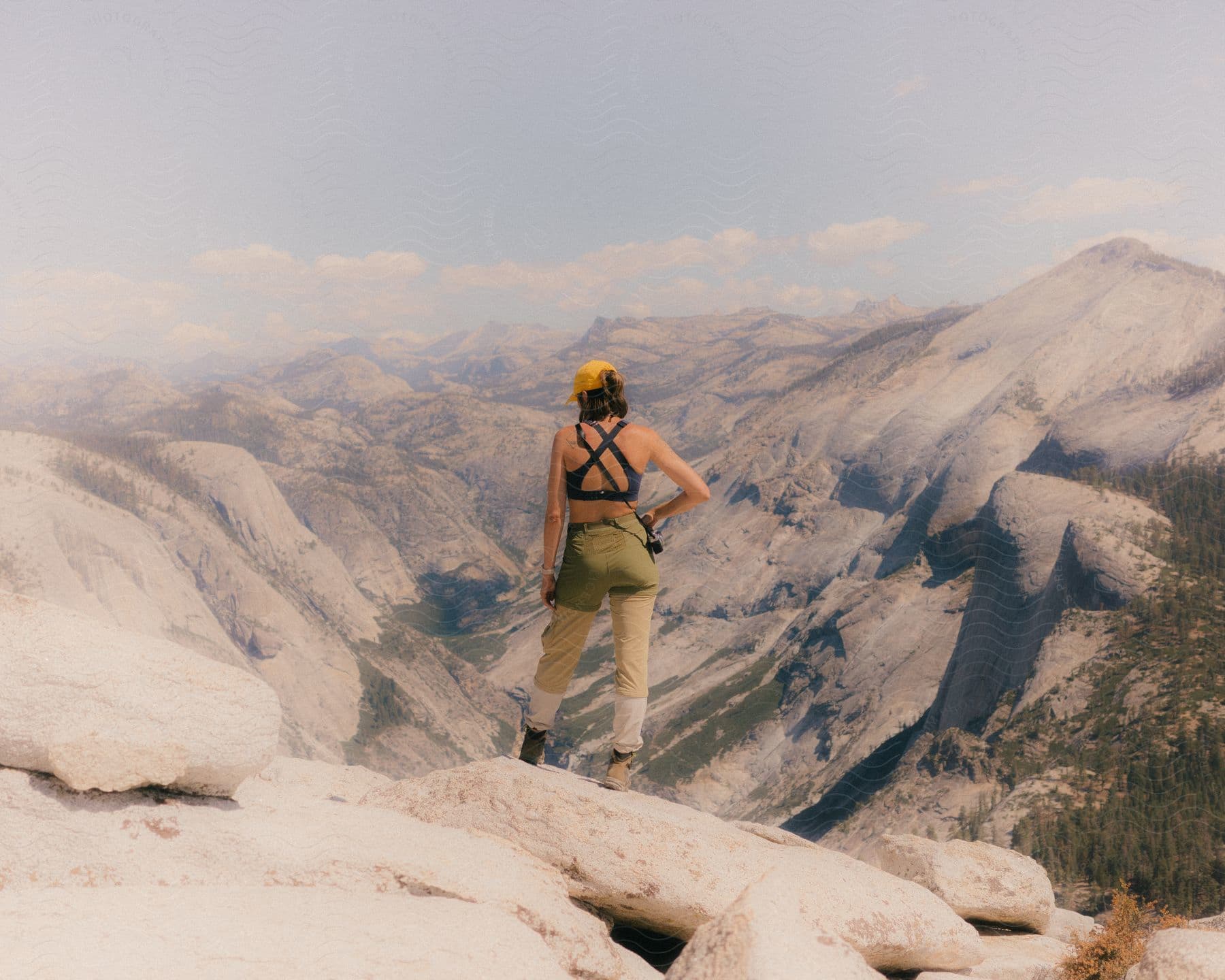 Woman wearing a yellow cap black top and green pants stands on top of mountain overlooking rocky valley