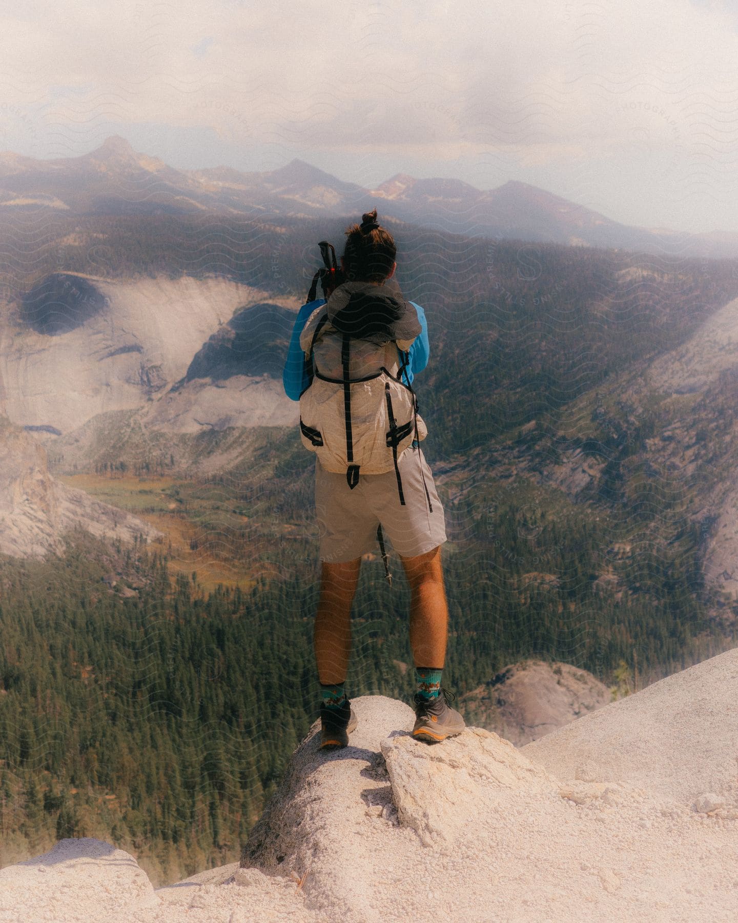 A male hiker with a backpack stands on a mountaintop, gazing at the valley forest below.