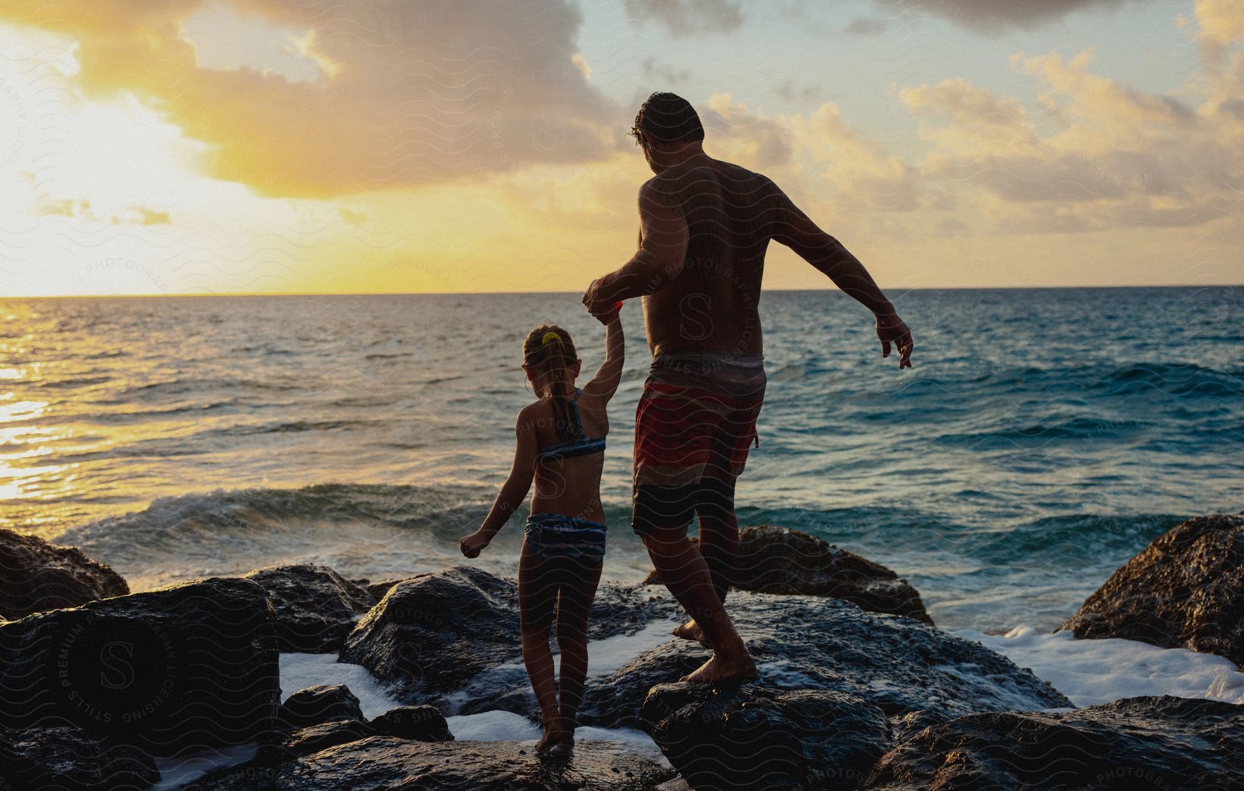 a man holds his daughter's hand while walking on rocks along the coast