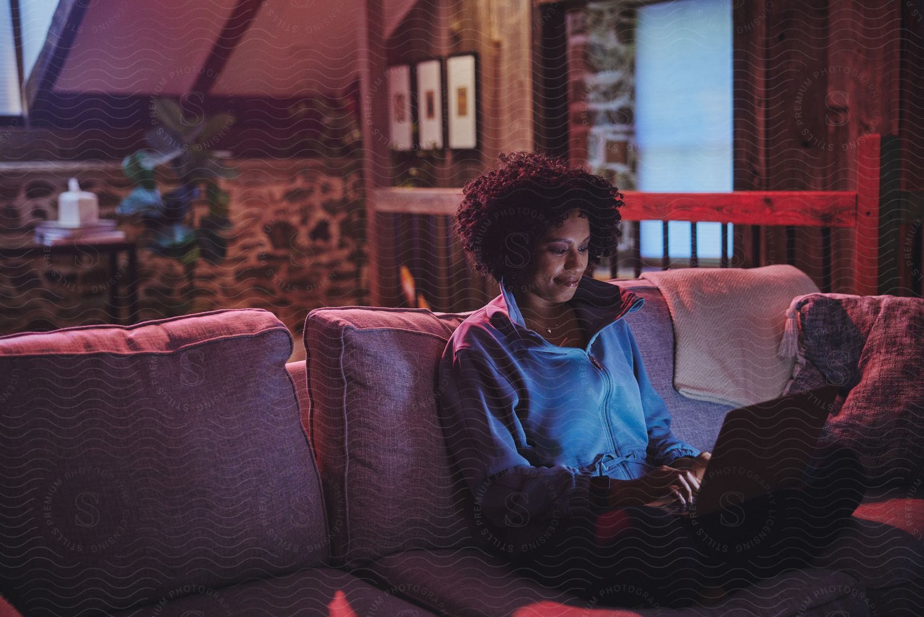 A woman in a blue sweater glows from her laptop screen as she sits on a couch in a rustic living room.