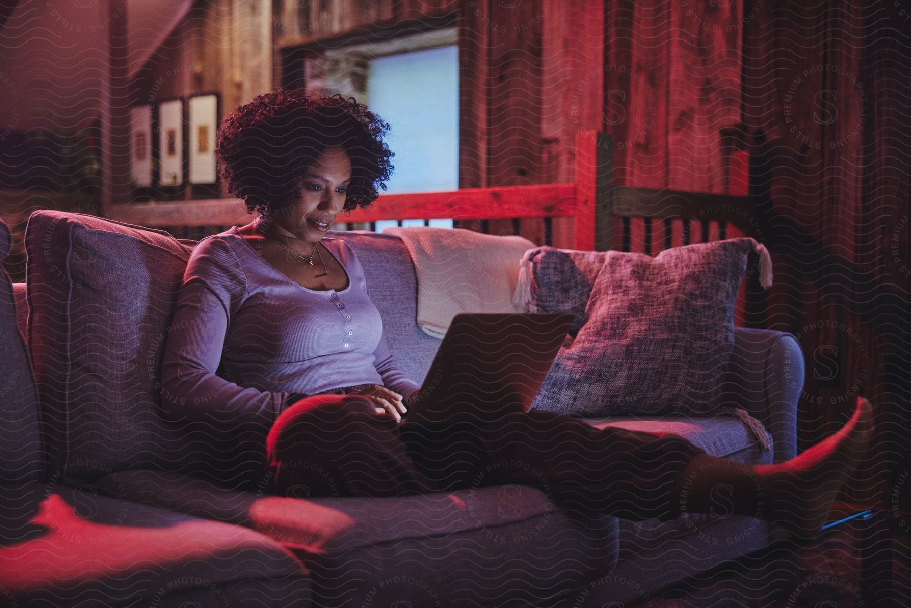 A woman with frizzy curly hair illuminated by red light sits on a couch at night, using a laptop.