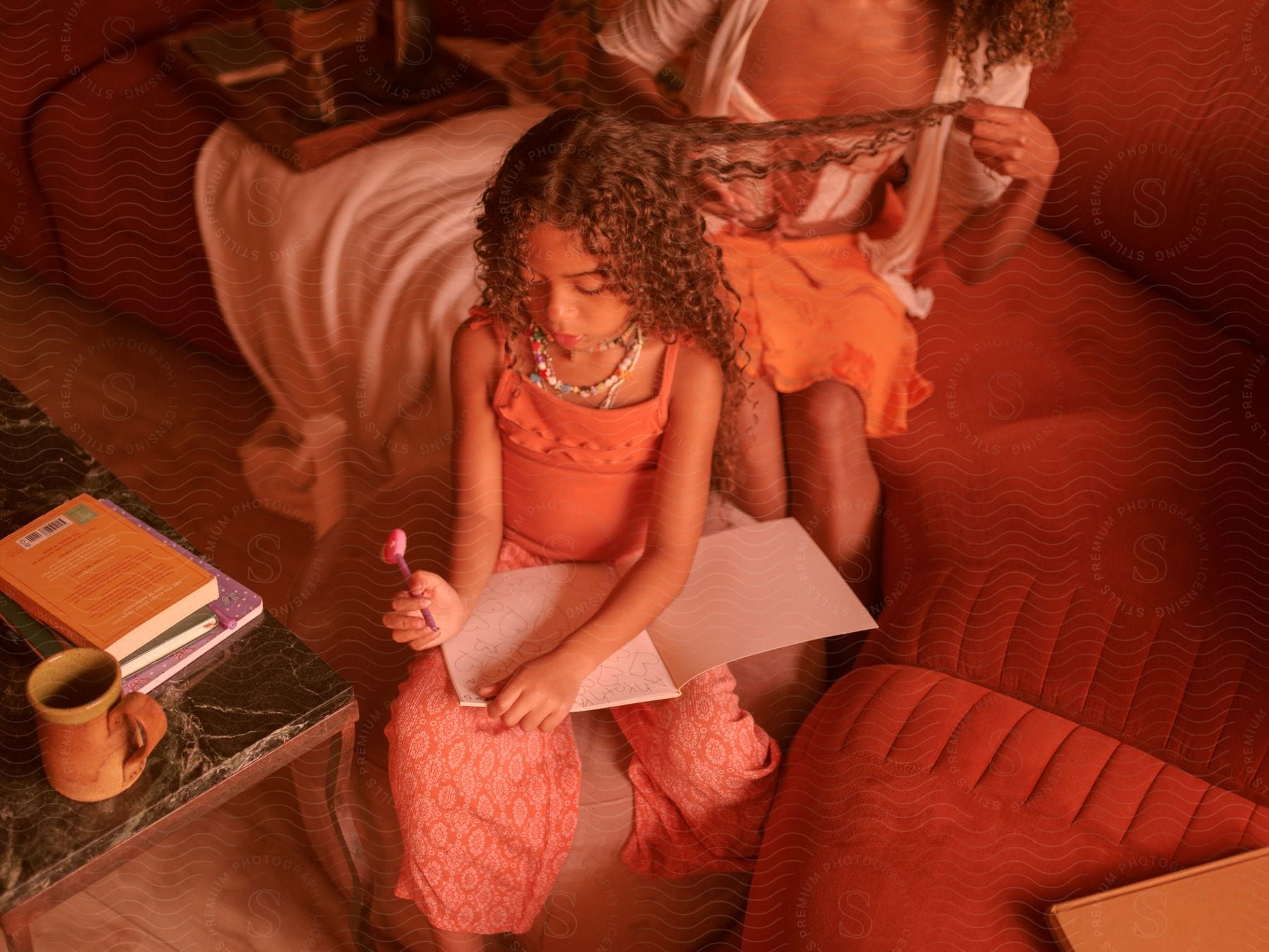 A child and her mother are sitting in the living room and the mother is doing the daughters hair while she draws in her notebook.