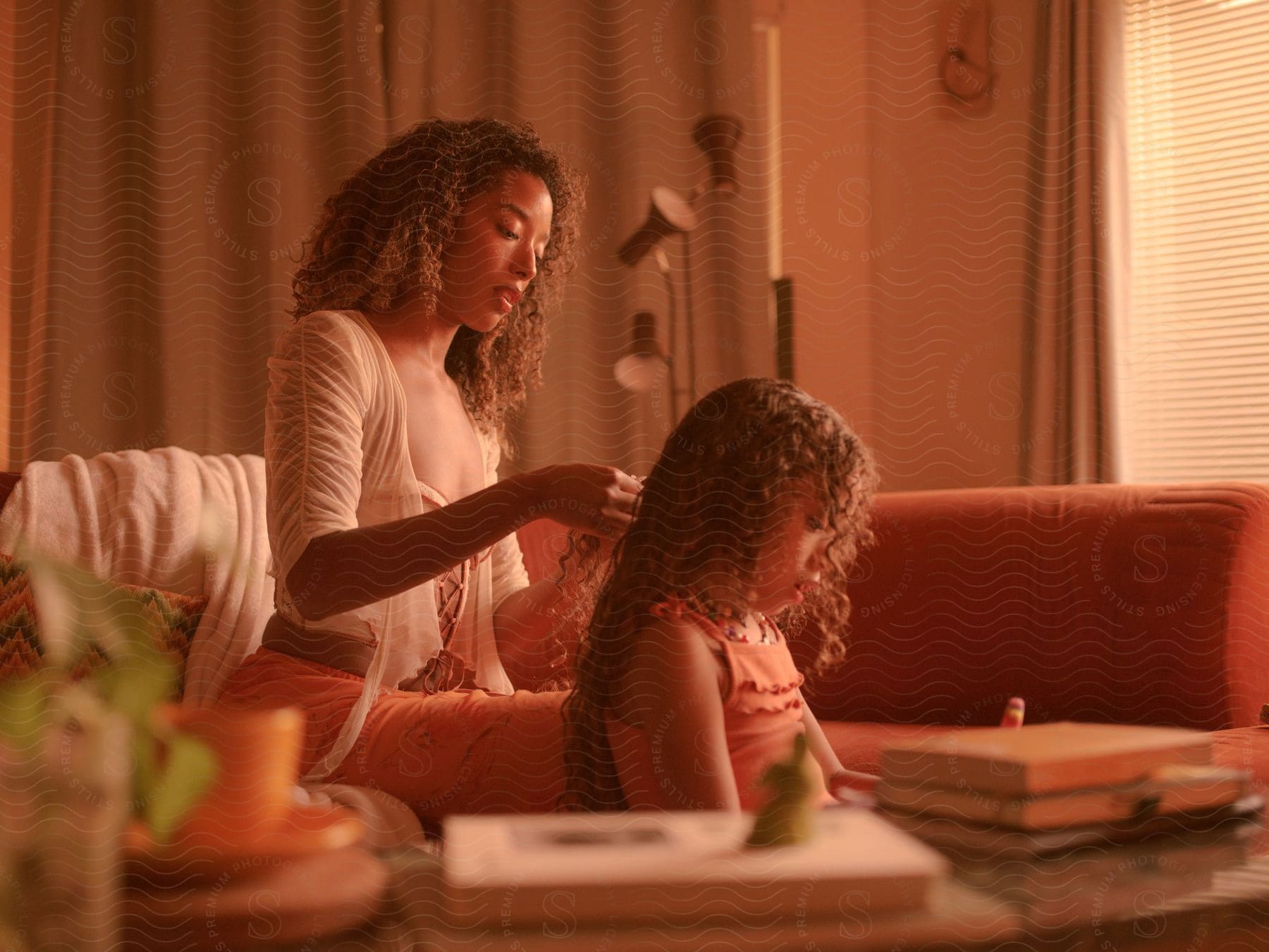 A mother making her daughter's hair while sitting on the couch indoor at mid-day