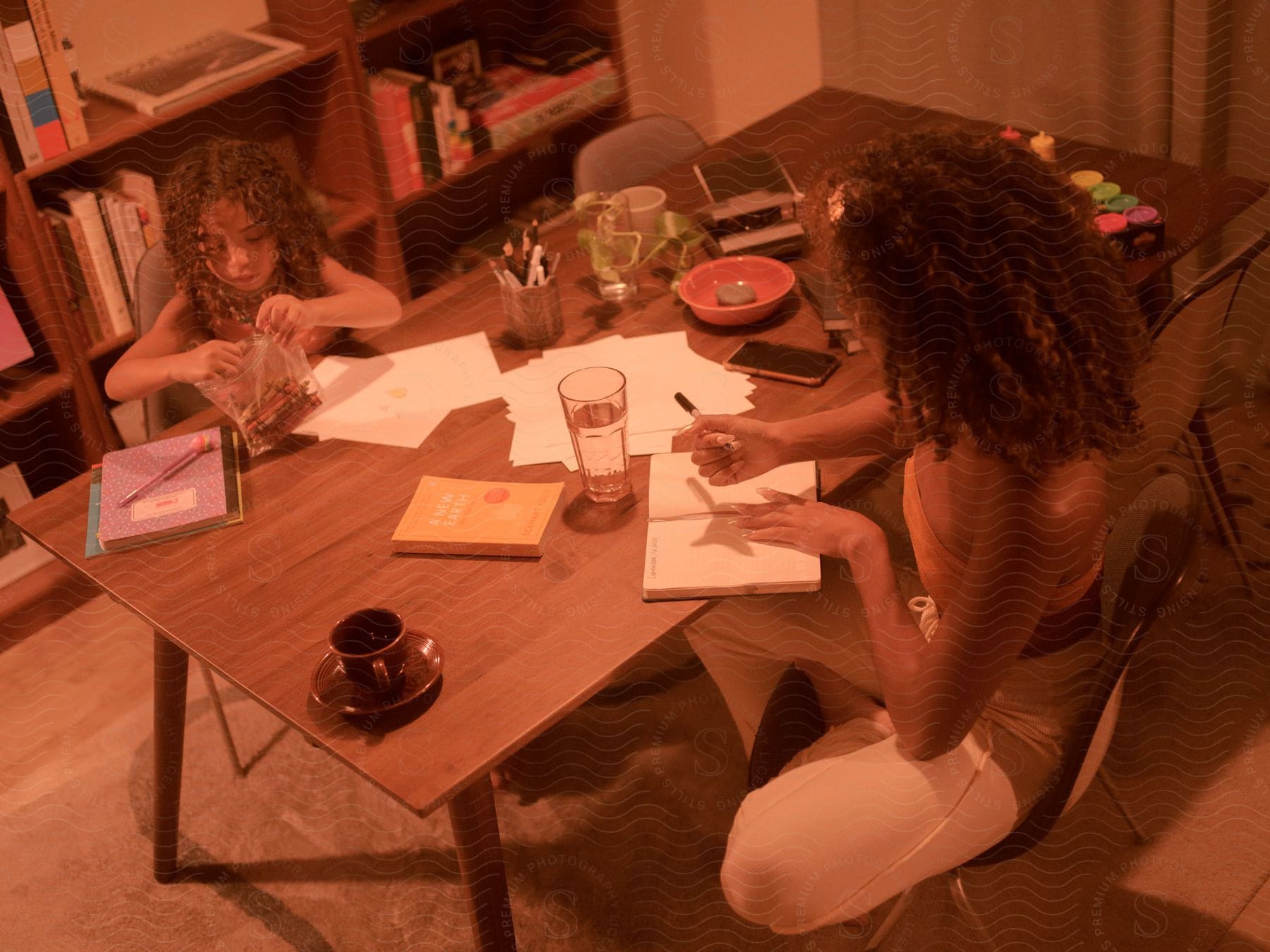 A girl with curly hair is looking through a plastic of crayons as a lady with curly hair is about to write something on a book. They are sitting on the table that has papers, other books, stationaries, cups, and bowls.