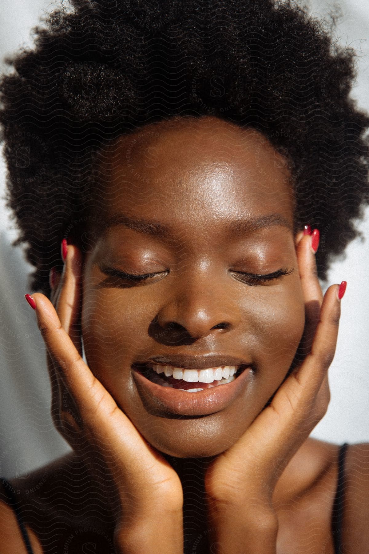 A radiant black woman with frizzy hair and red fingernails smiles with her hands to her face, her eyes closed.