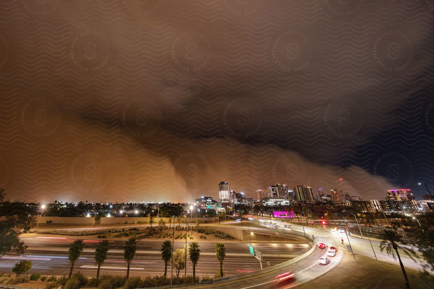 Traffic moves on the highways and lights on towers and buildings glow in the distance as dark storm clouds pour rain over the city