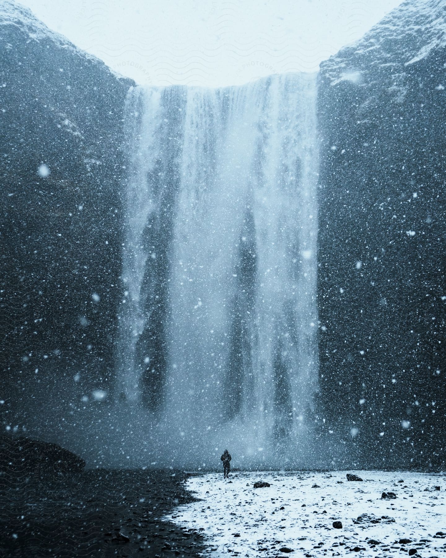 Snow fills the air as a man walks on ice as waterfalls flow over the cliff