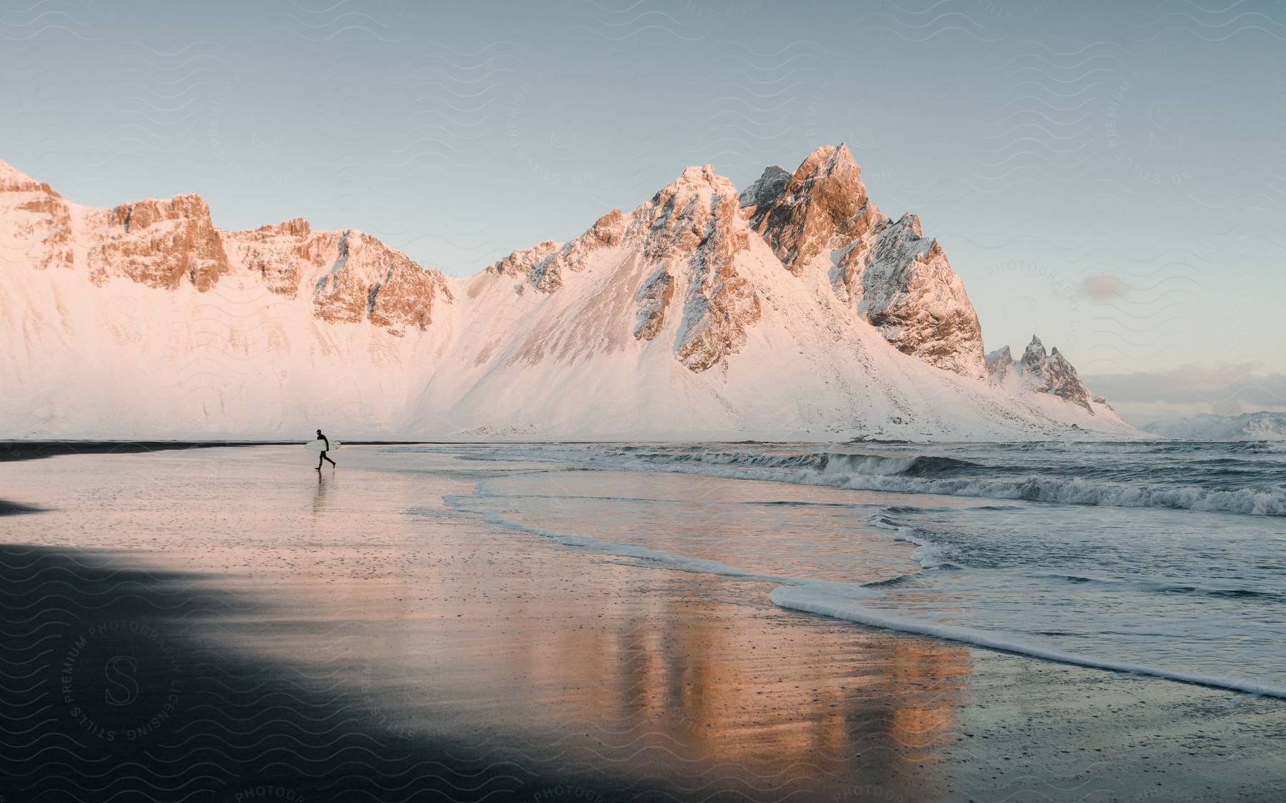 A surfer on the sand of a beach with snow-capped mountains in the background.