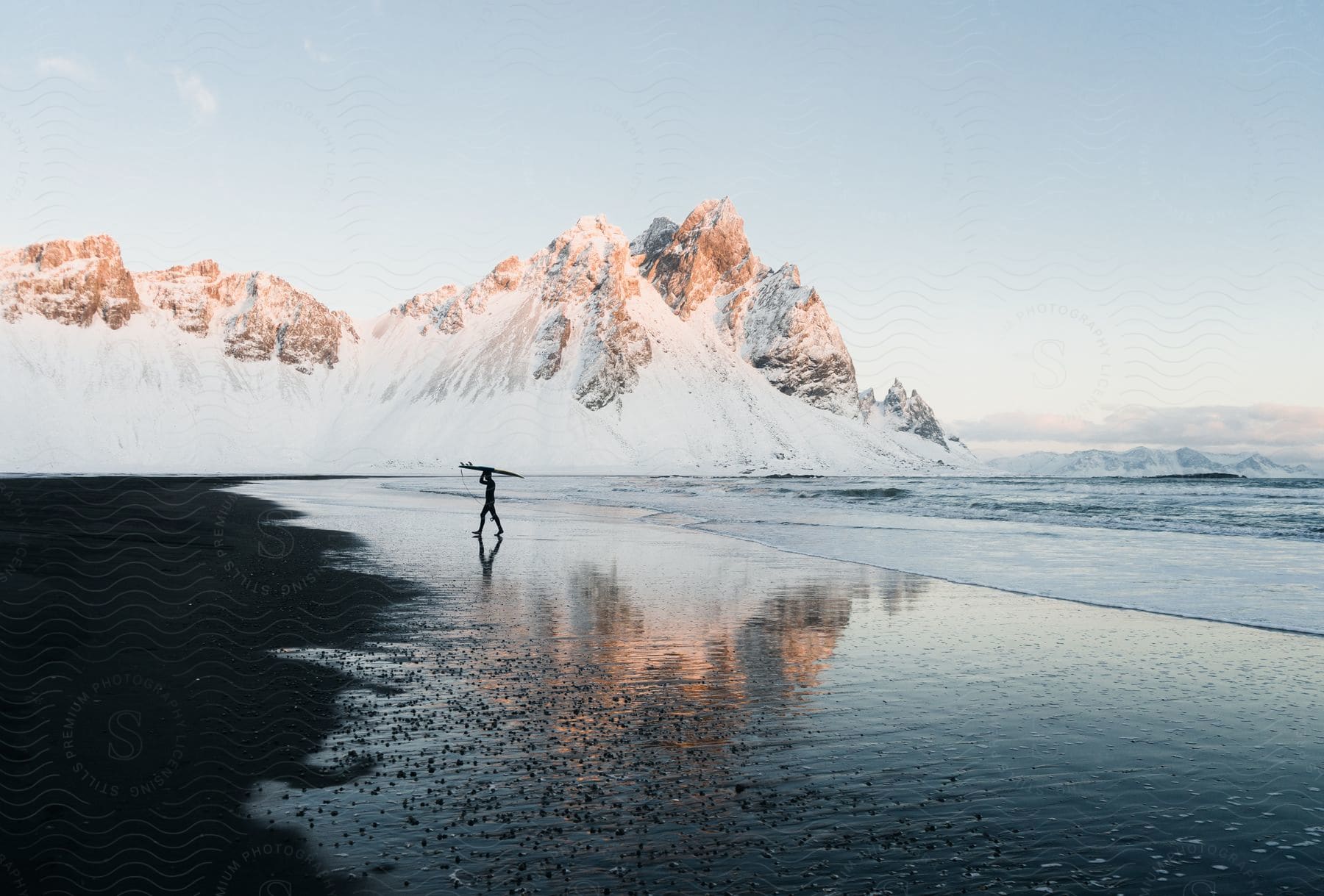 Lone Surfer In Silhouette Carries Surfboard While Crossing Wet Beach Sand That Reflects Nearby Snowy Mountains