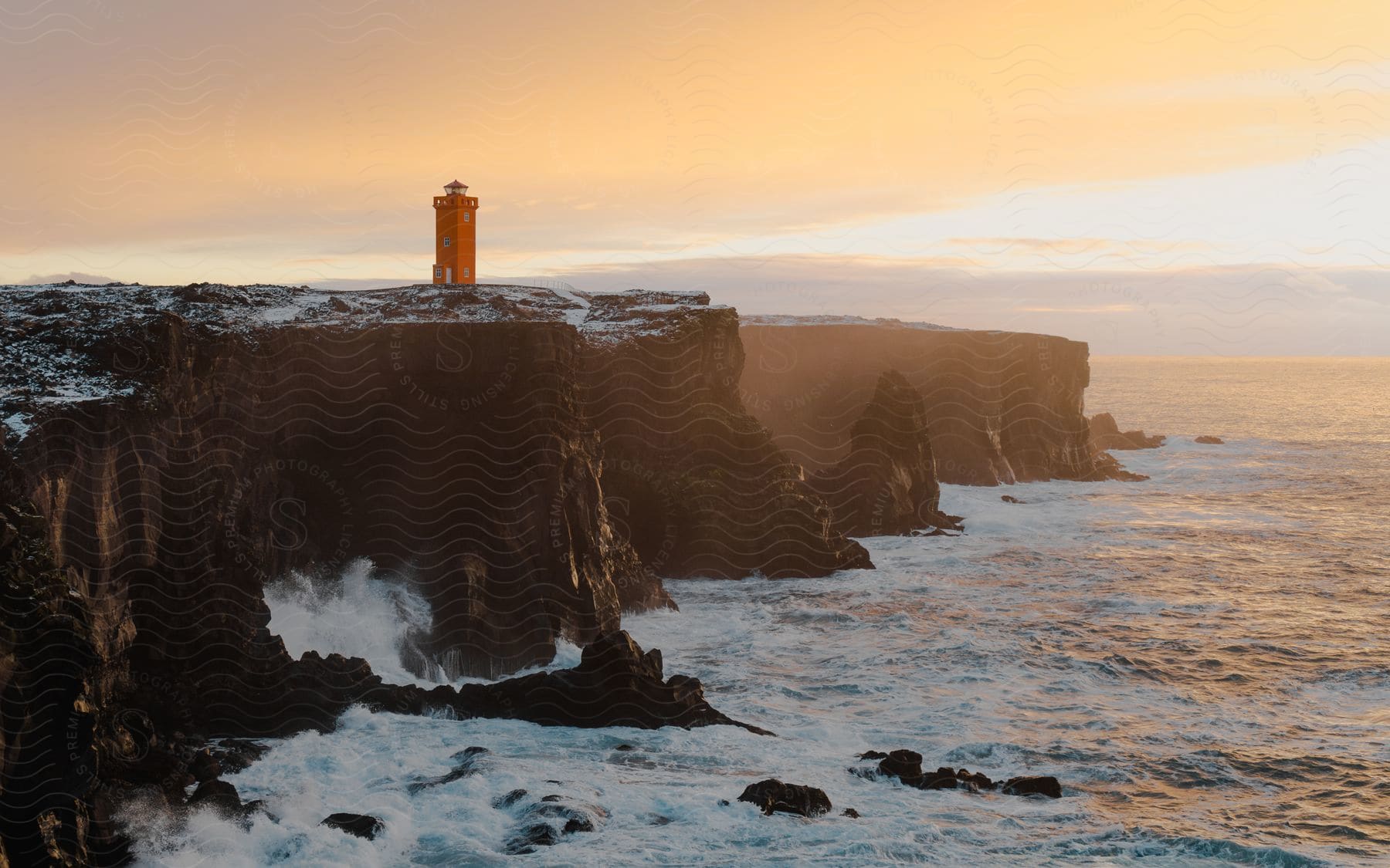 A lighthouse sits at the edge of a coastal cliff line at sunset.