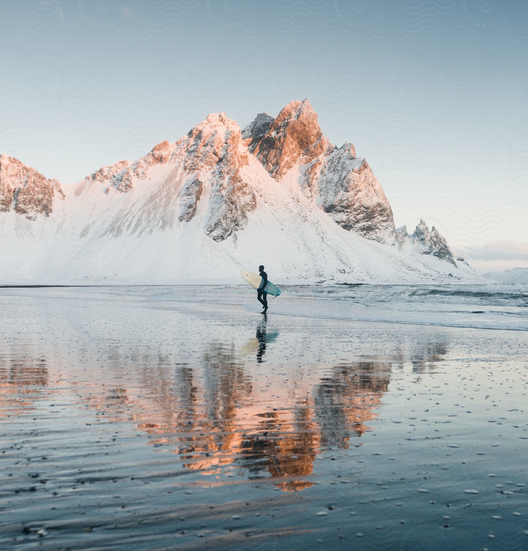 Surfer carries surfboard across reflective sand near snow covered mountain.