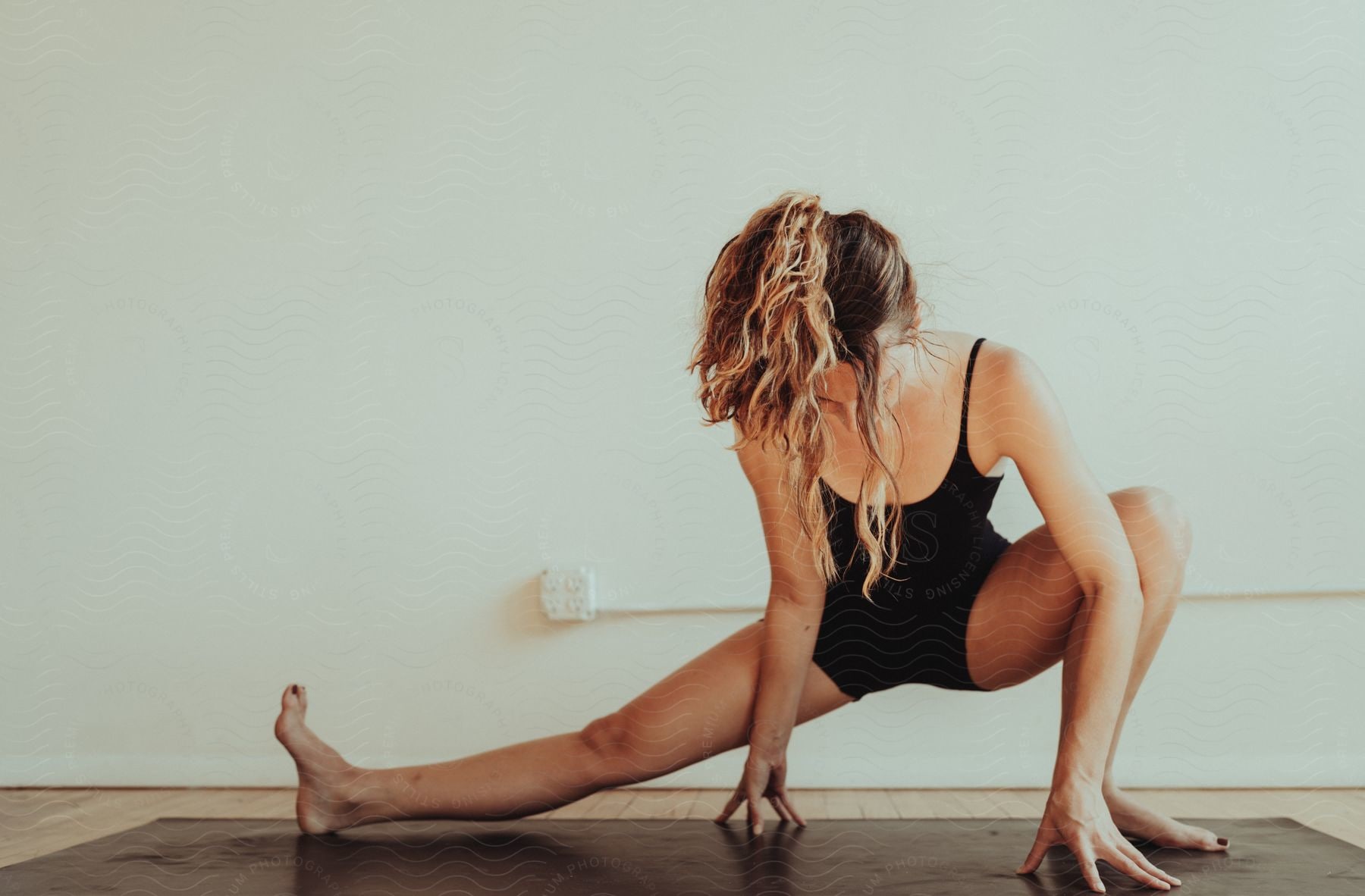 A woman in shorts performing a yoga pose, stretching her right leg to the side while bending her left knee and placing both hands on the yoga mat on the floor.